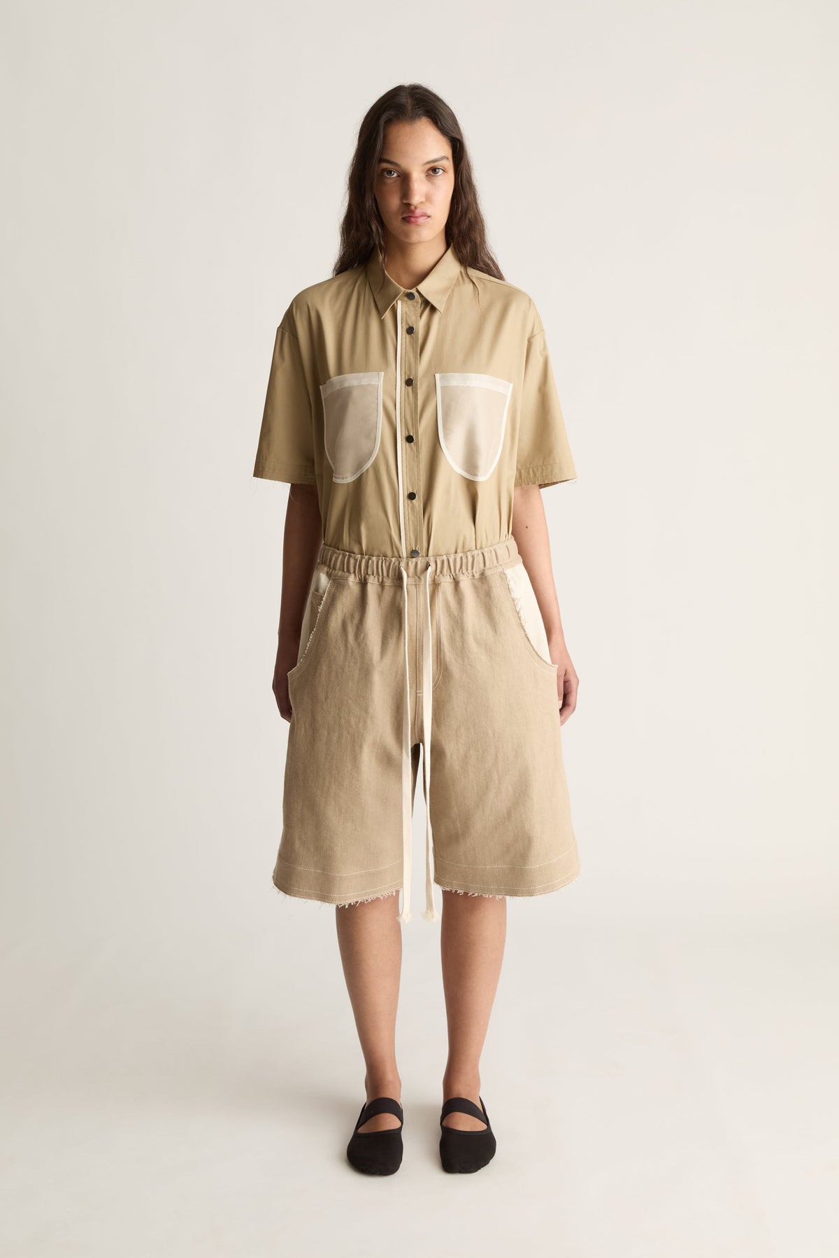 Front view of a woman wearing a beige short-sleeve button-up shirt and drawstring bermuda shorts with cream pockets against a white studio background