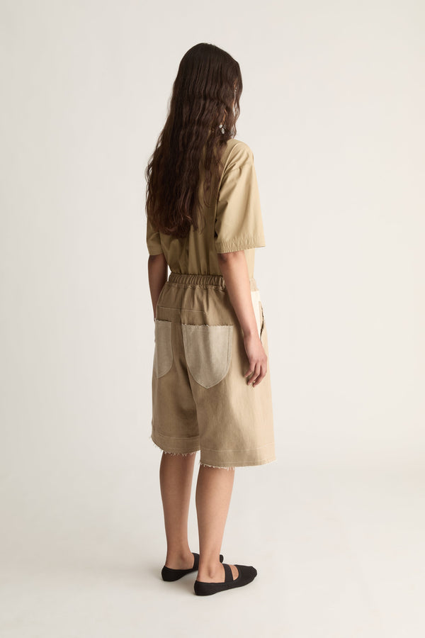Back view of a woman with long wavy hair in a beige short-sleeve shirt and bermuda shorts with cream back pockets