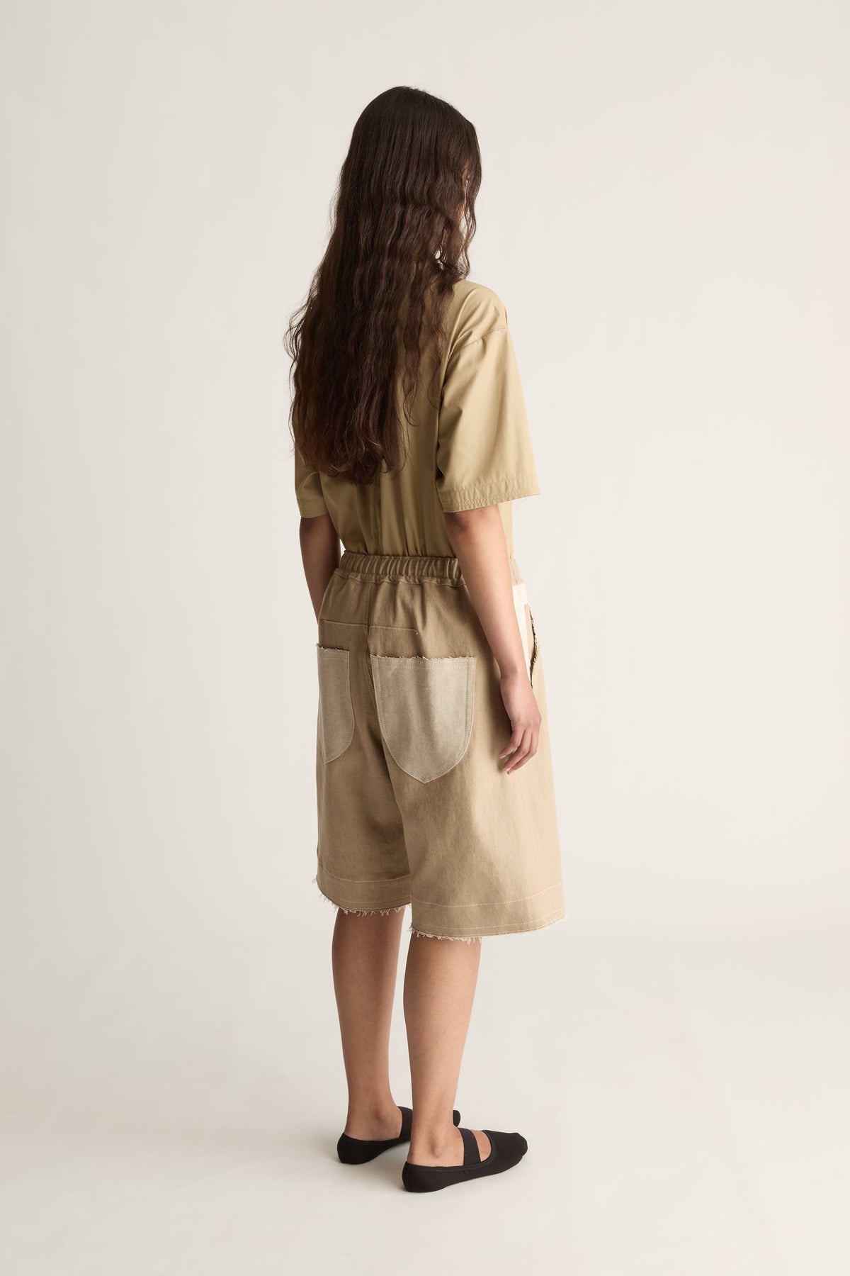 Rear view of a woman with long wavy hair in a matching beige short-sleeve top and knee-length shorts with black ballet flats