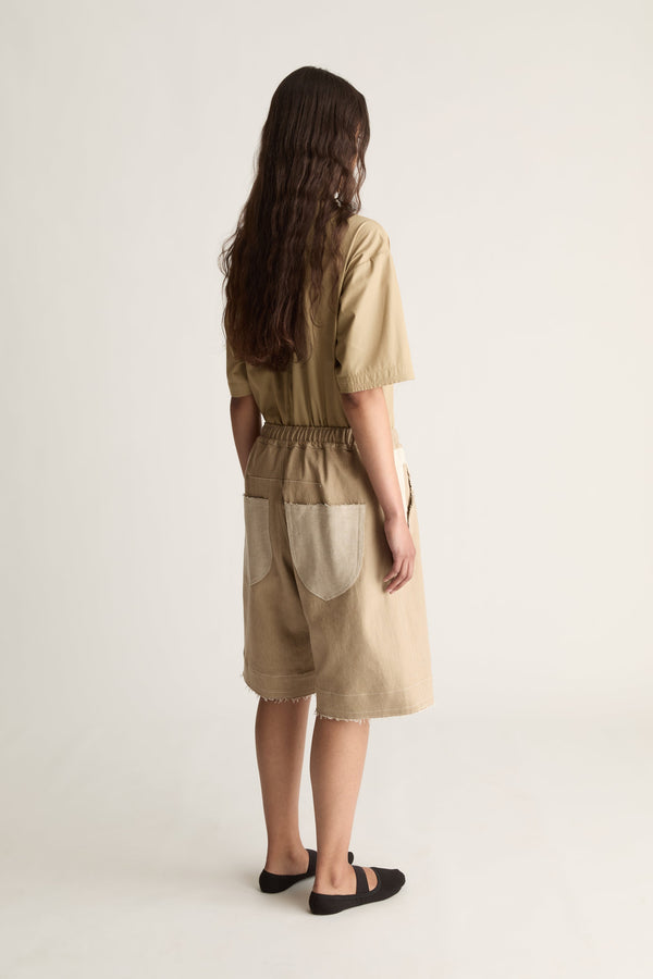 Rear view of a woman with long wavy hair in a matching beige short-sleeve top and knee-length shorts with black ballet flats