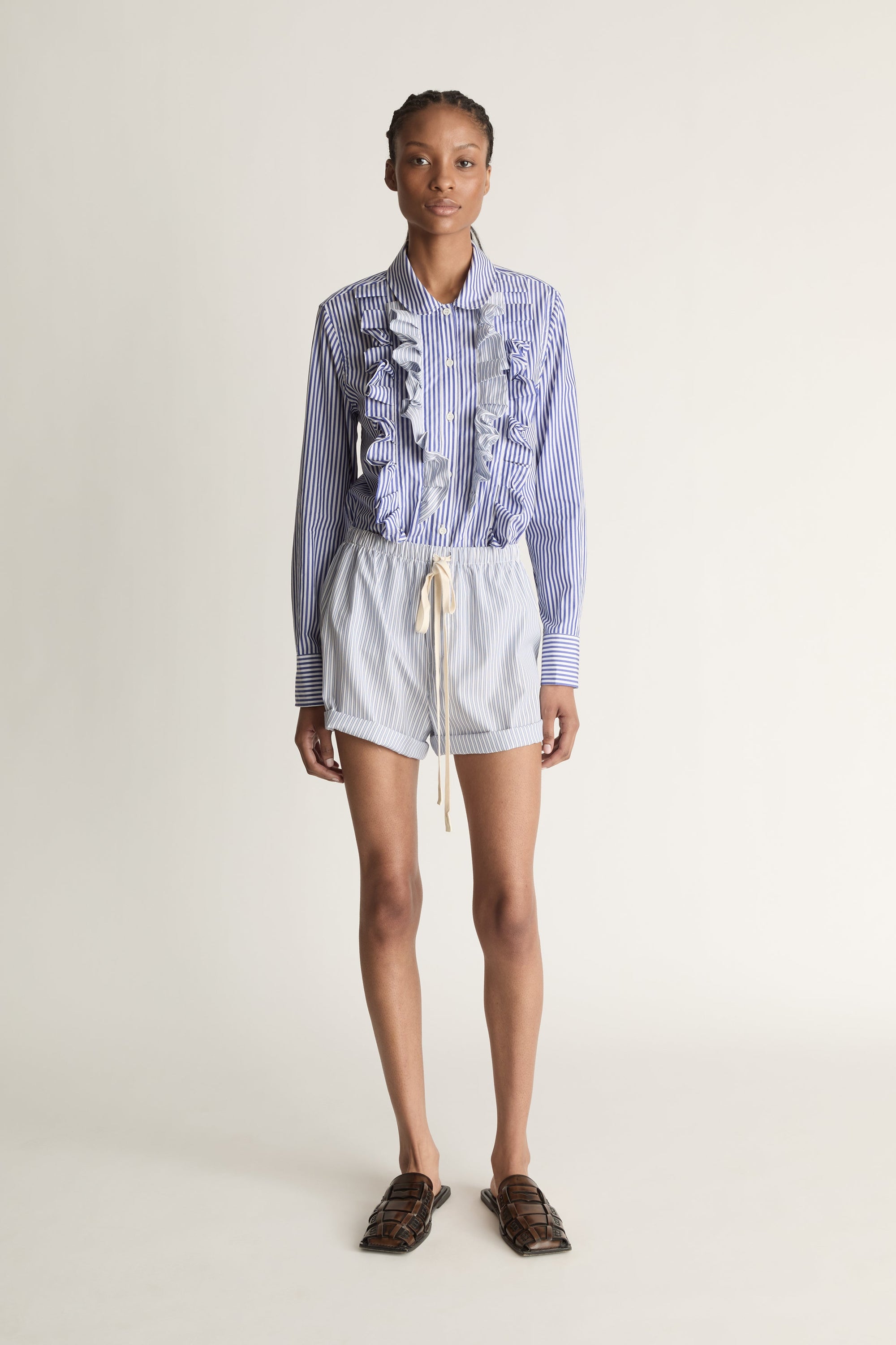 Person wearing a blue and white striped shirt with ruffled details and white shorts on a plain background