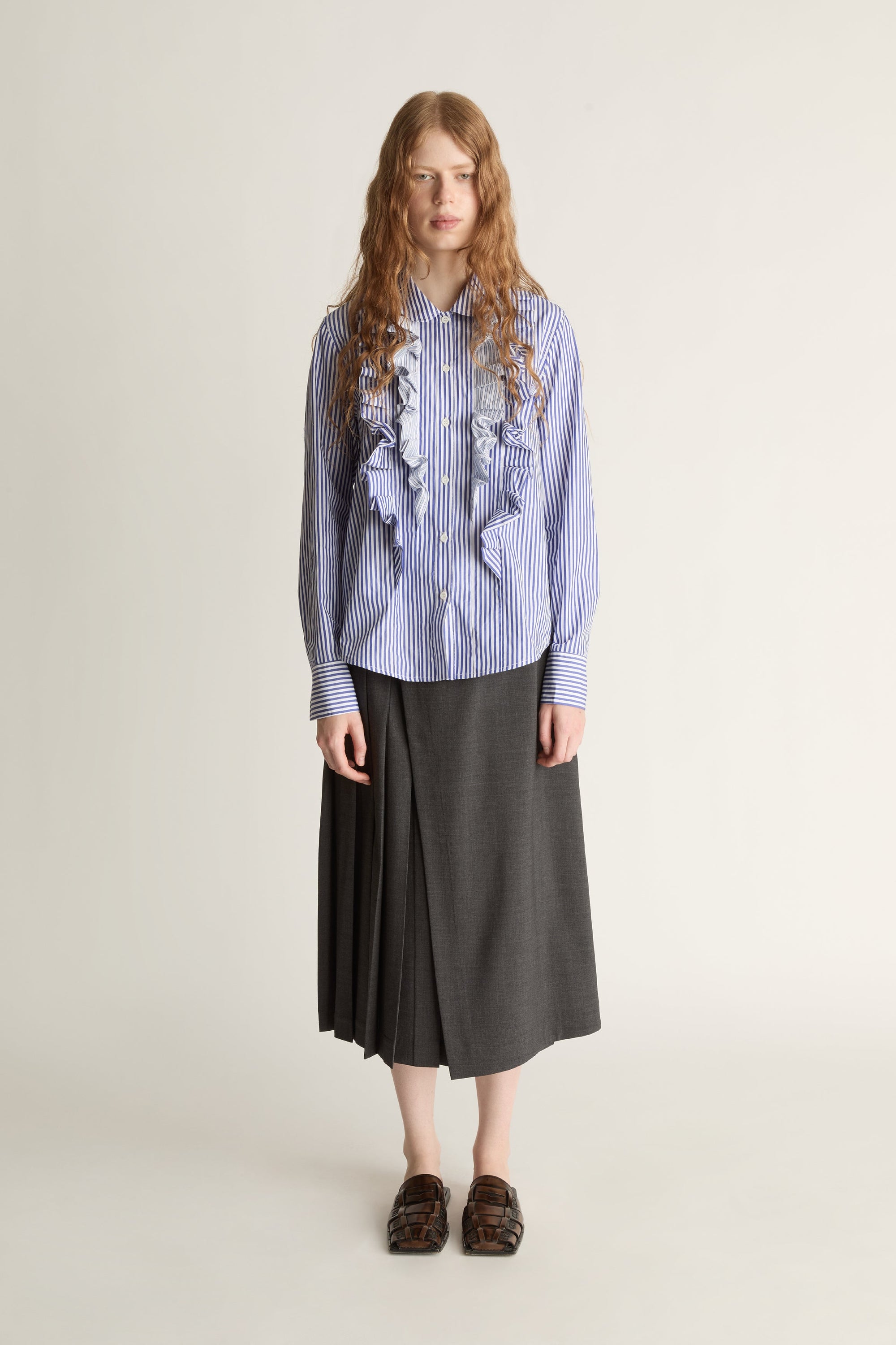 Woman wearing a blue striped shirt with ruffles and a gray skirt on a white background