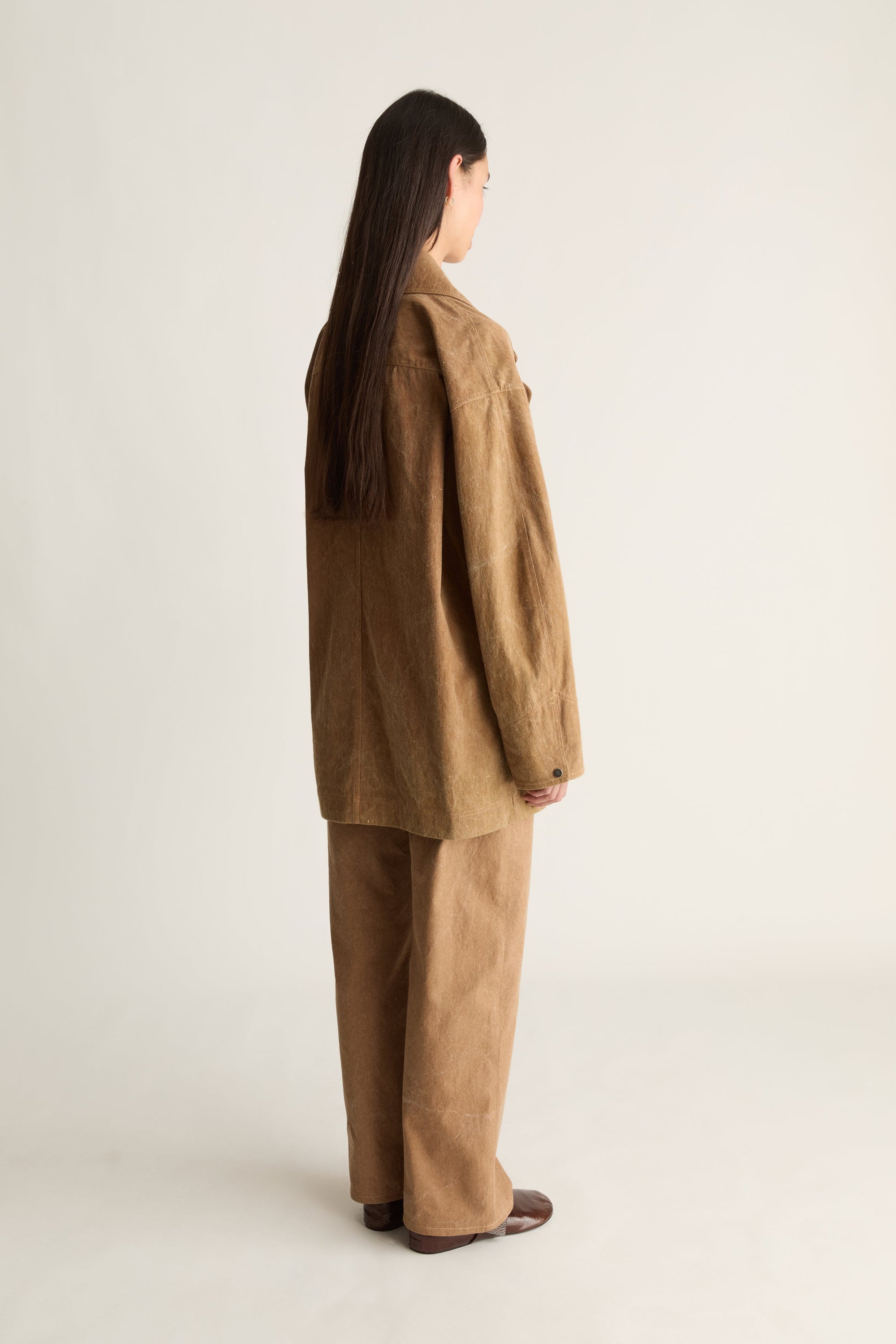 Woman wearing a brown coat and pants on a white background, facing the back.