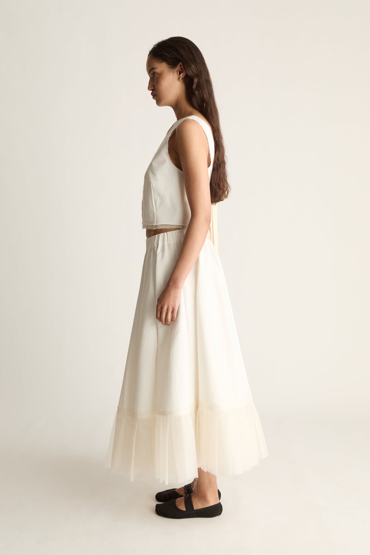 Side view of a full cream maxi skirt flaring out to a sheer layered hem, worn with a cream cropped top.