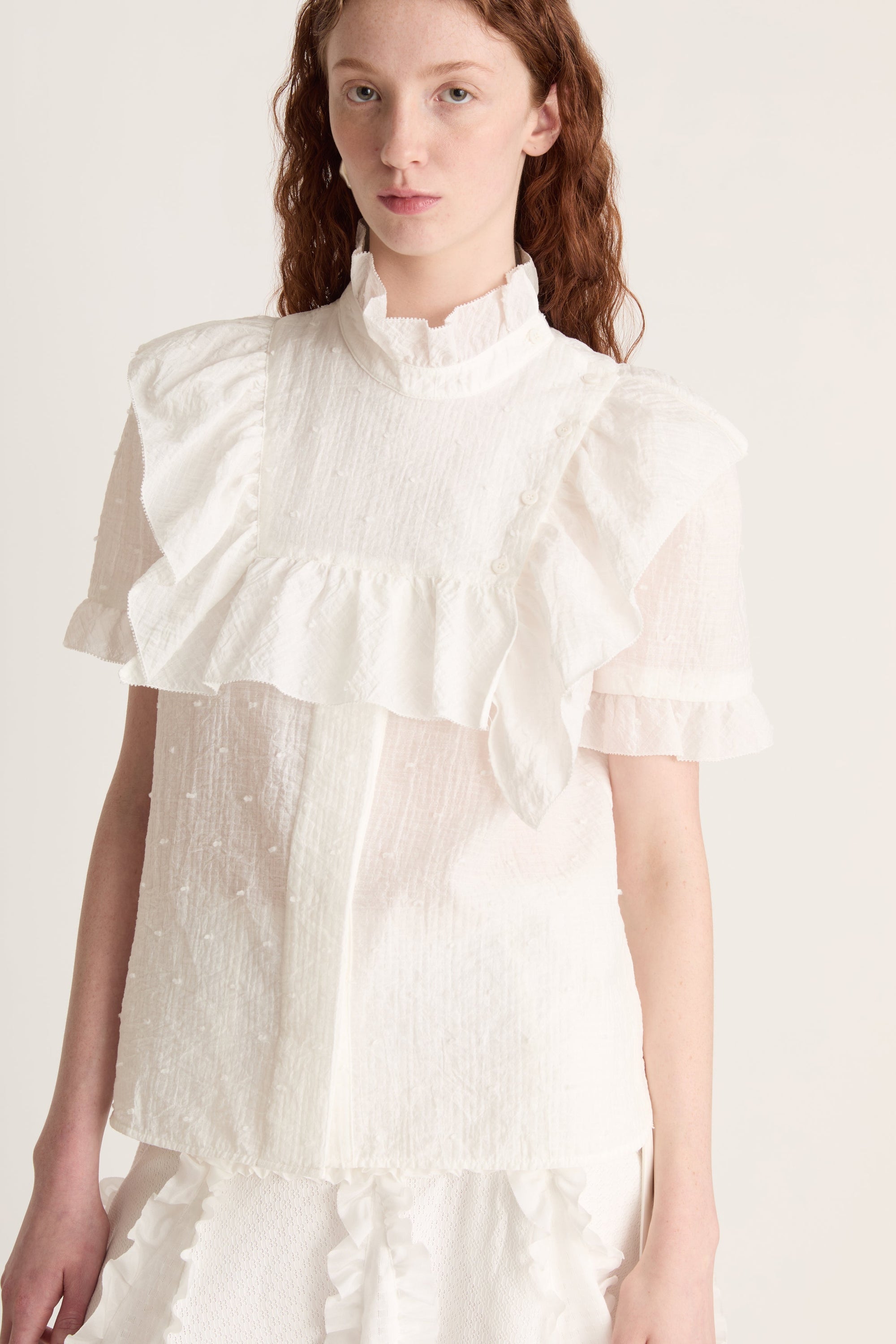 A woman wearing a white top with ruffled sleeves  and a white skirt on a plain background