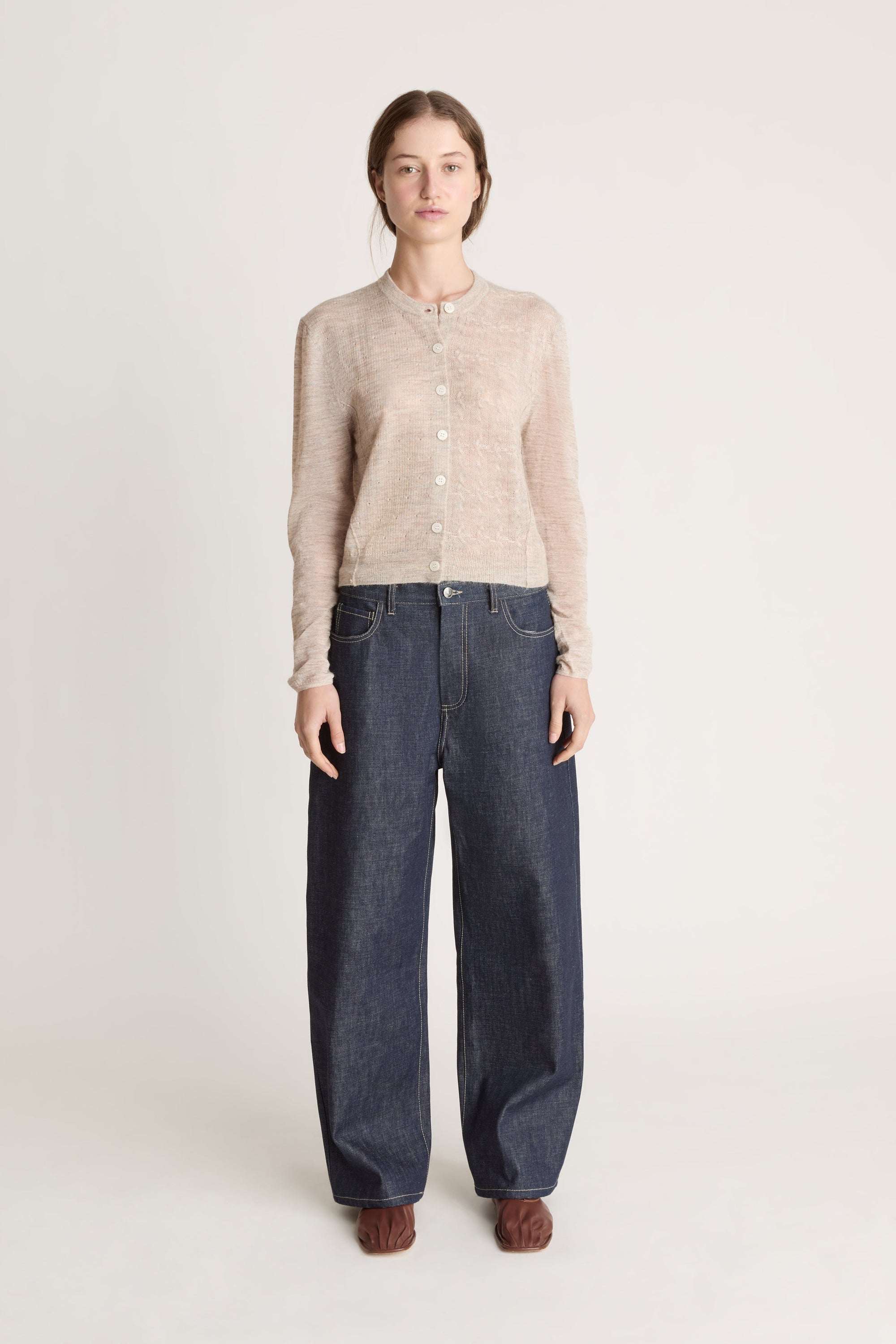 Person wearing a beige cardigan and dark jeans on a white background