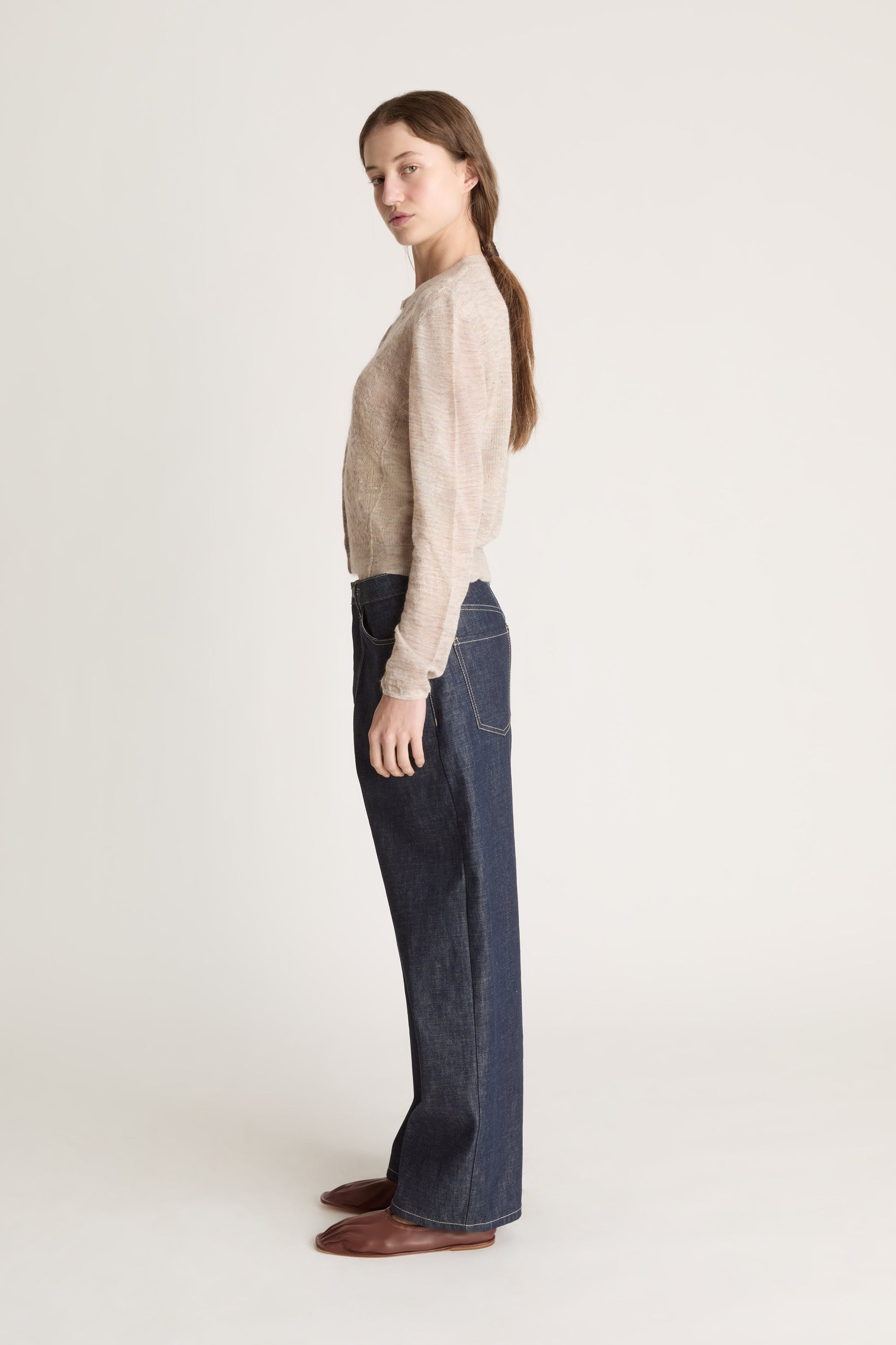 Person wearing a beige cardigan and dark jeans on a white background