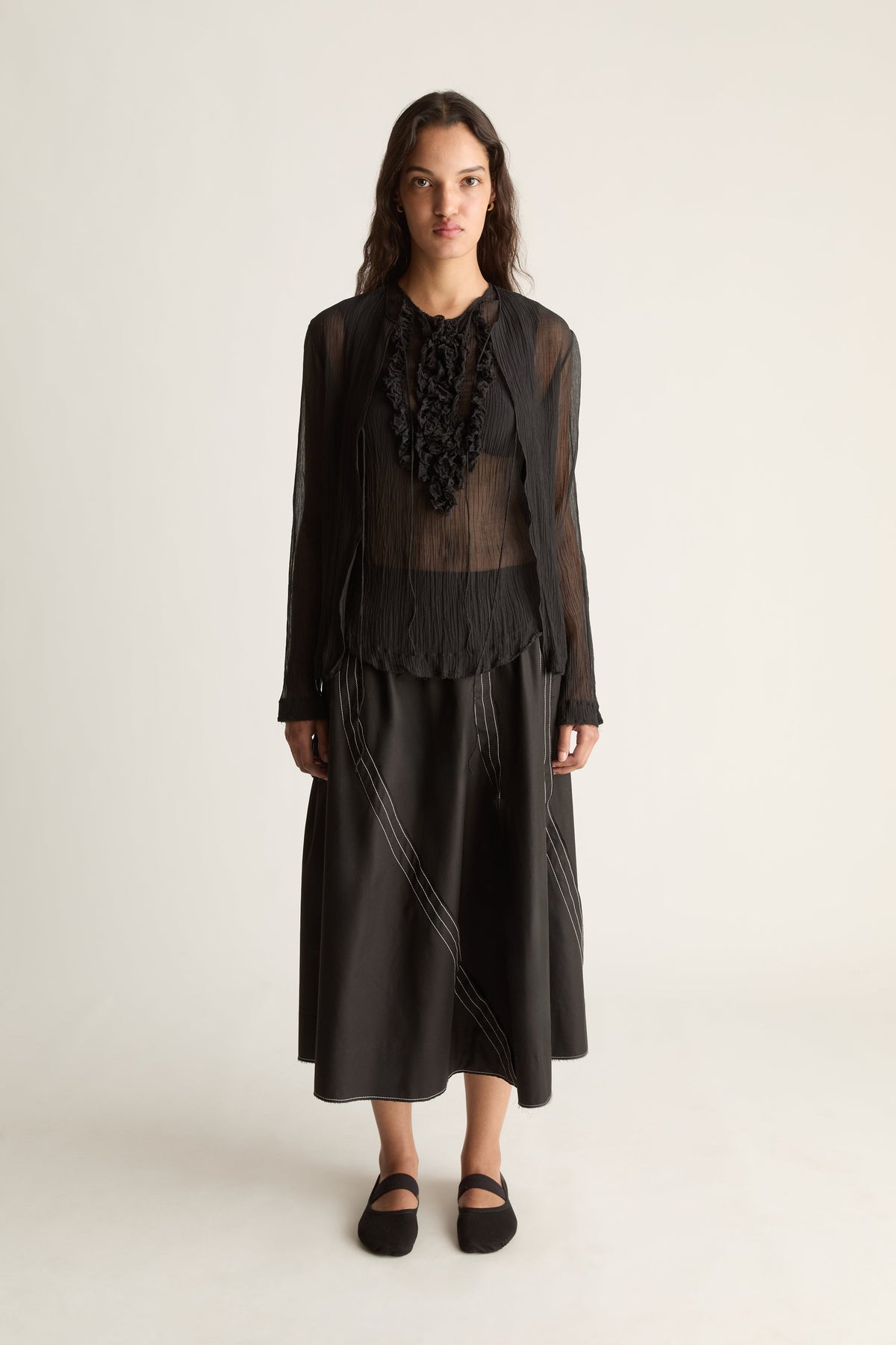 Front view of a woman wearing a sheer black open-front cardigan over a ruffle top and black midi skirt against a white studio background
