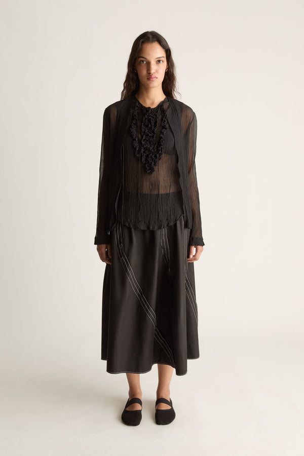 Front view of a woman wearing a sheer black open-front cardigan over a ruffle top and black midi skirt against a white studio background