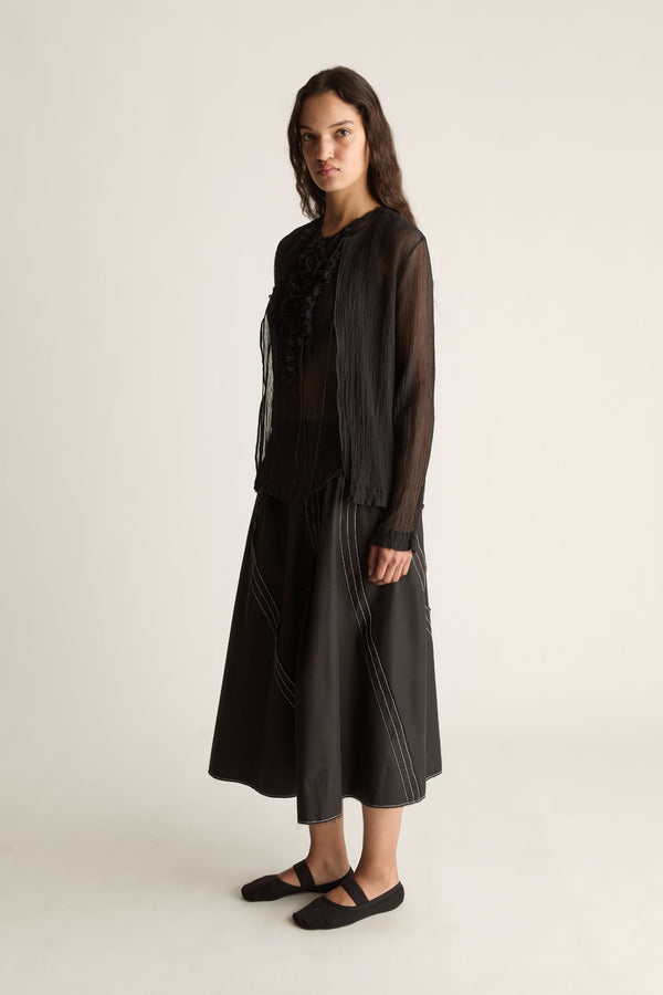 Angled view of a woman in a long-sleeve sheer black cardigan, ruffle top and black skirt with white stitching