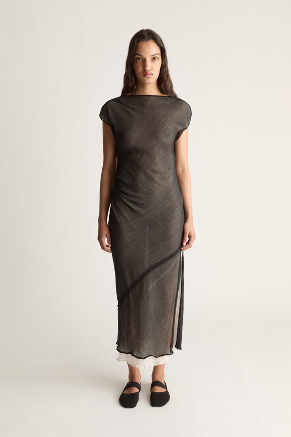 Front view of a woman wearing a sheer black boatneck midi dress with cap sleeves and ivory underlayer against a white studio background