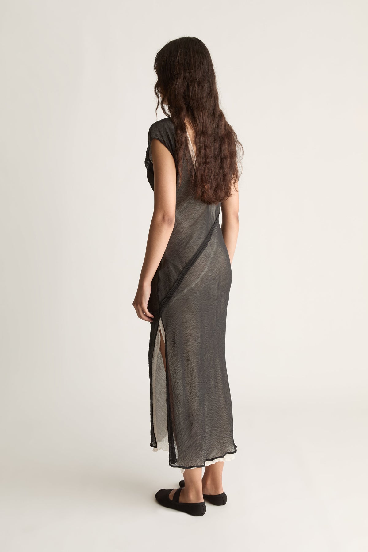 Back view of a woman with long wavy hair in a sheer black maxi dress with ivory slip and diagonal seam