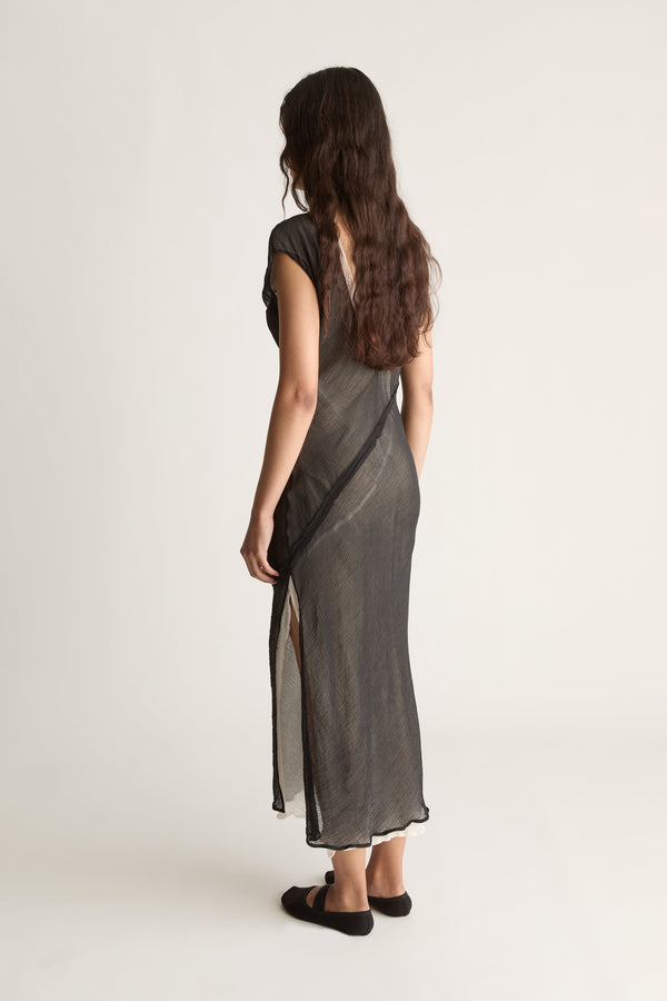 Back view of a woman with long wavy hair in a sheer black maxi dress with ivory slip and diagonal seam