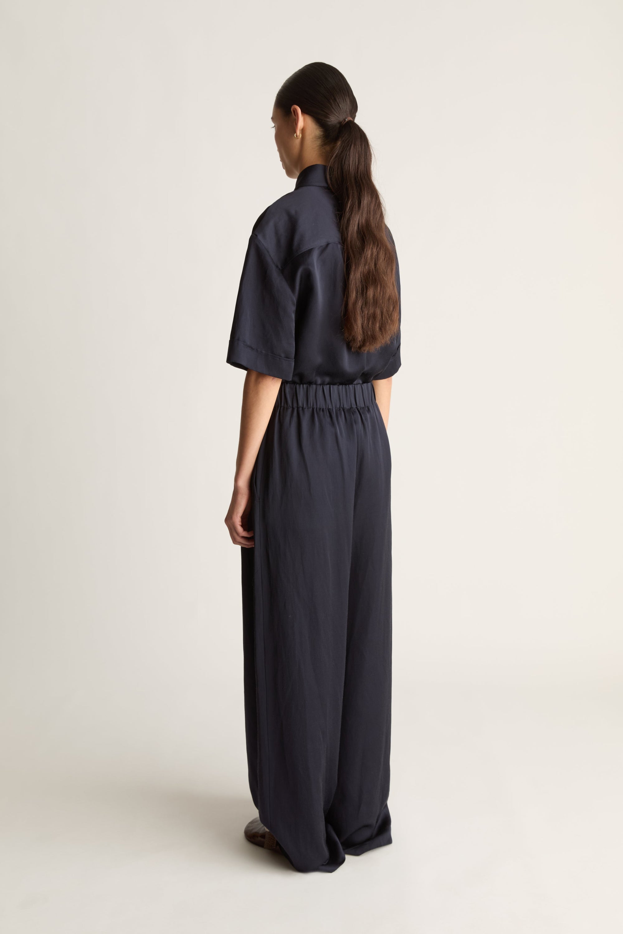 A model shown from the back wearing a dark navy blue short-sleeve collared top and matching wide-leg trousers, photographed against a plain light gray studio background.