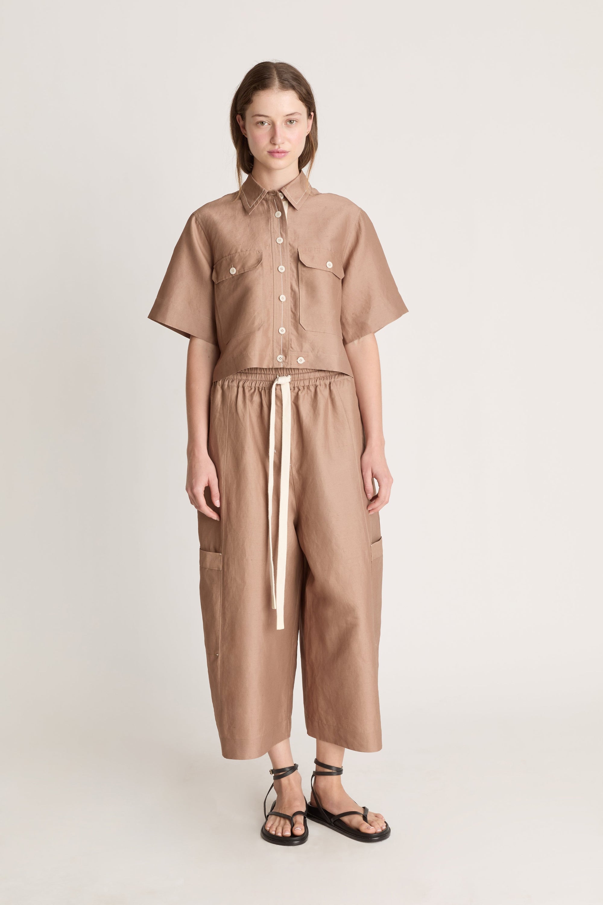 Woman wearing a brown button up short sleeve shirt and brown pants on a white background 
