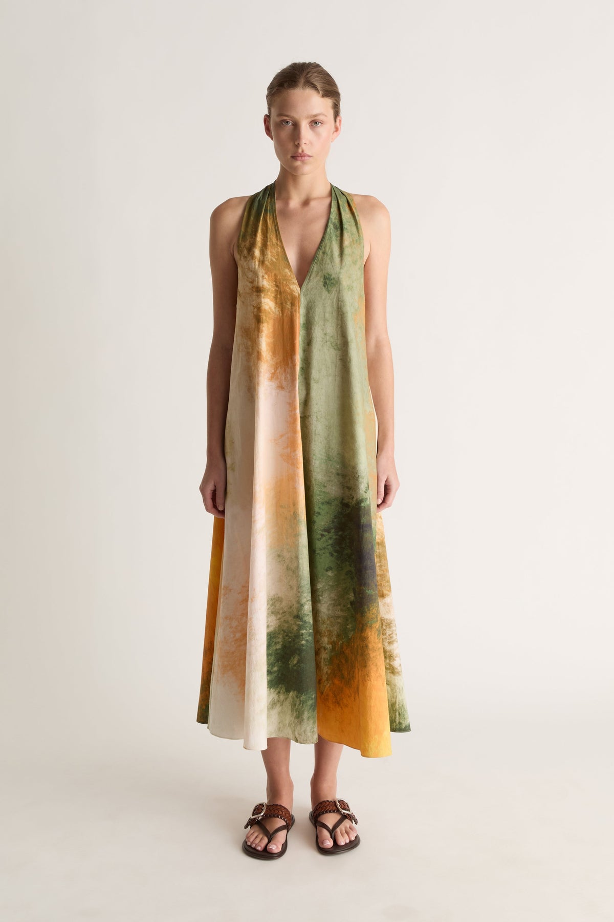Front view of a woman wearing a green and orange watercolour-print halter midi dress with brown sandals against a white studio background