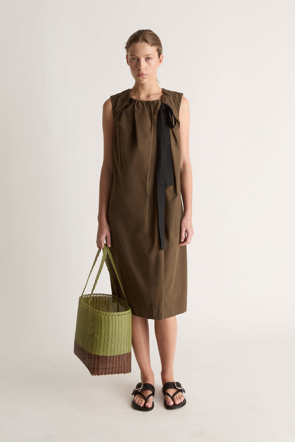 Front view of a woman in an olive-brown shift dress holding a green woven basket, with black sandals