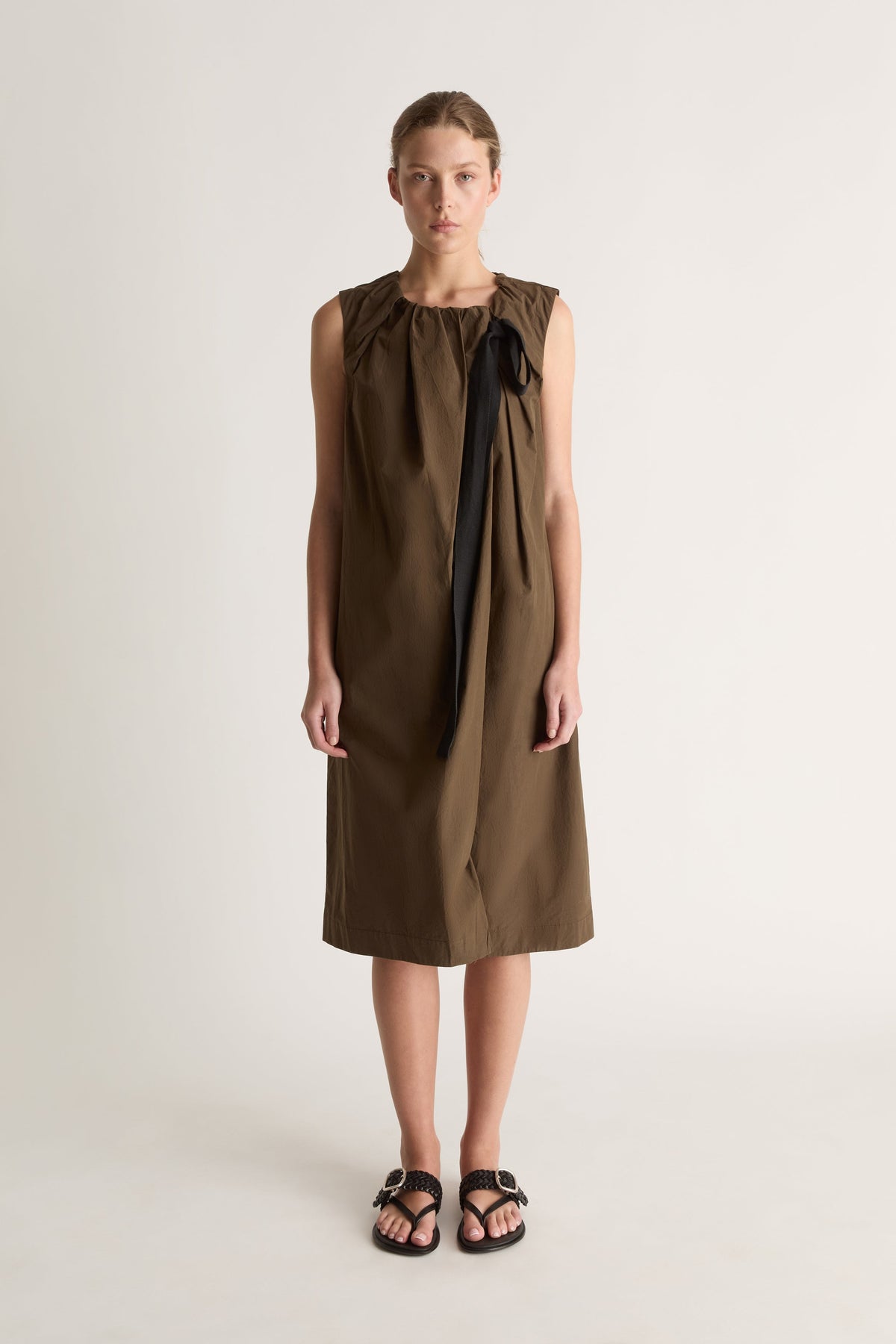 Front view of a woman wearing a khaki sleeveless midi dress with black ribbon tie against a white studio background