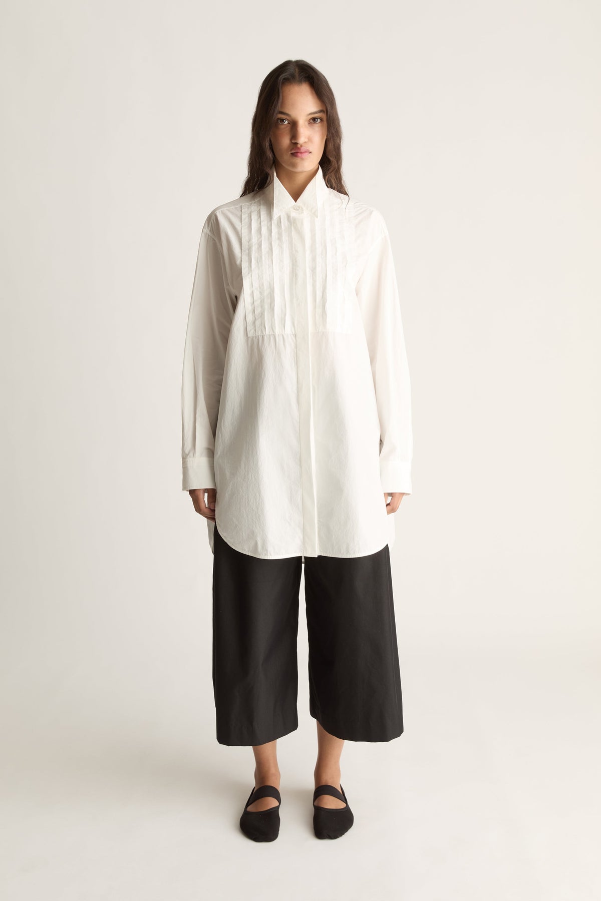 Front view of a woman wearing an oversized white tuxedo shirt with pleated bib and black cropped wide-leg pants against a white studio background