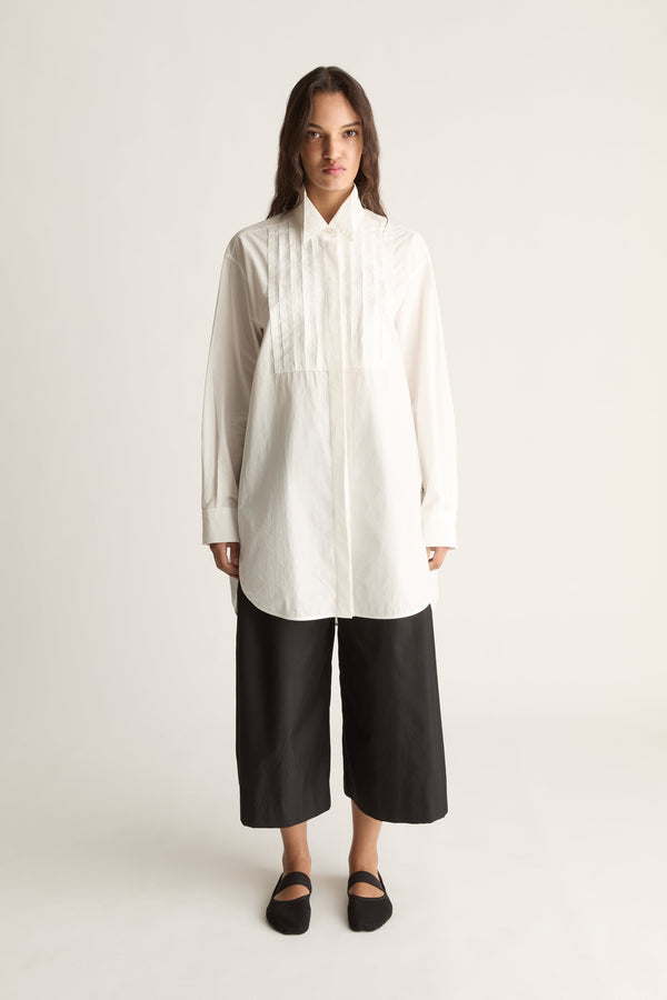 Front view of a woman wearing an oversized white tuxedo shirt with pleated bib and black cropped wide-leg pants against a white studio background
