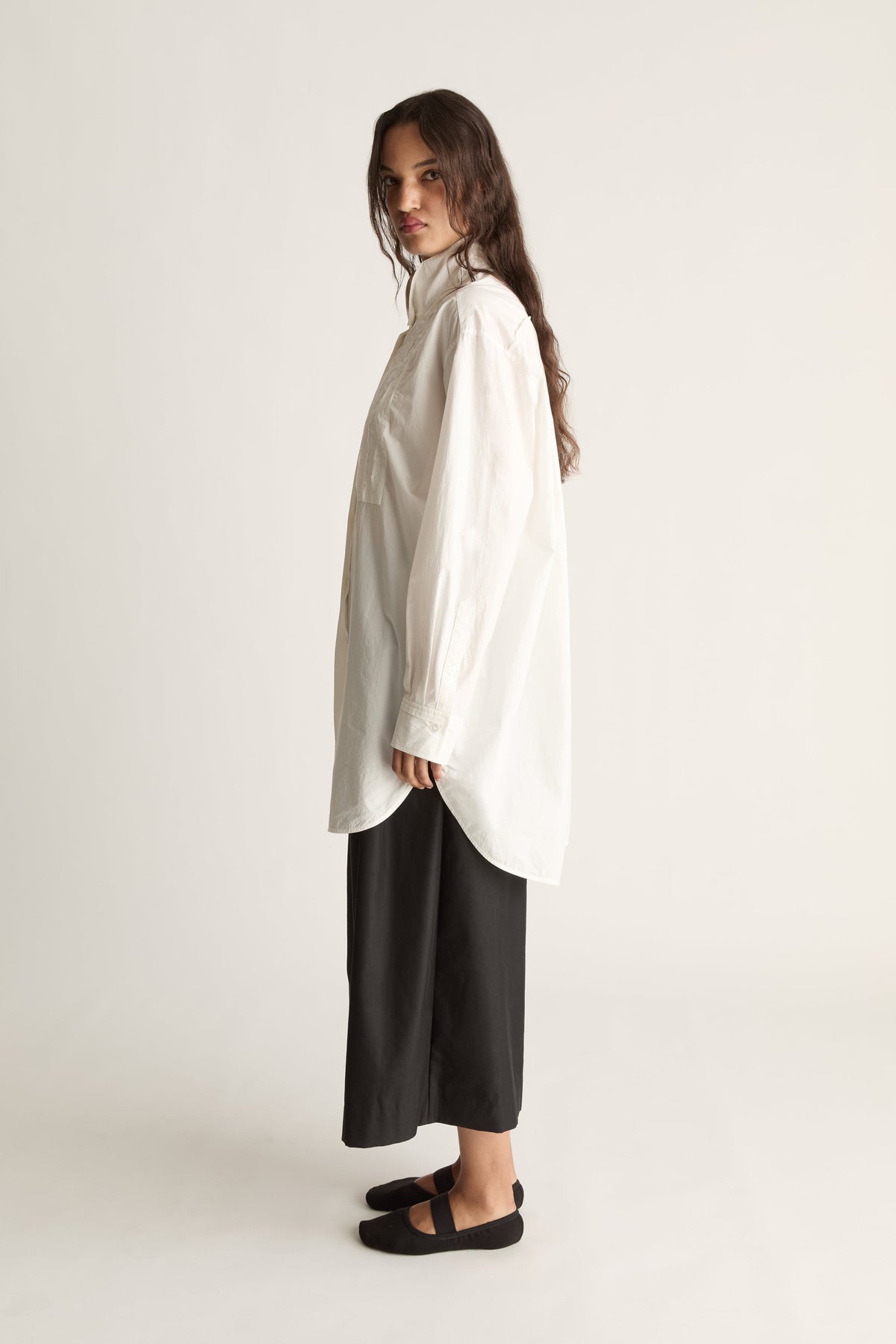 Side view of a woman in an oversized white long-sleeve shirt with chest pocket, black cropped pants and black ballet flats
