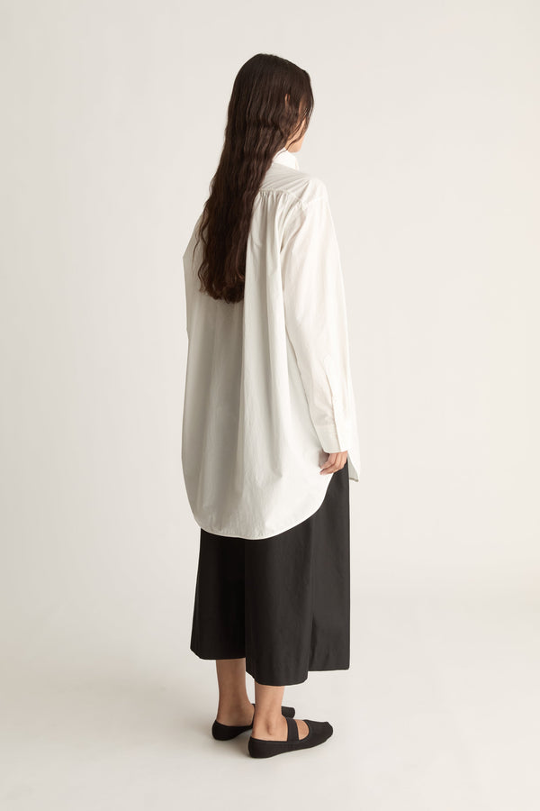 Back view of a woman with long dark hair in an oversized white shirt with curved hem and black wide-leg cropped pants