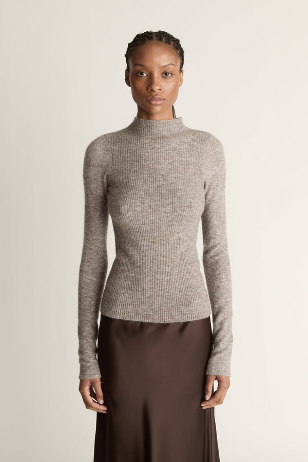 Woman wearing a gray turtleneck sweater and brown skirt against a beige background