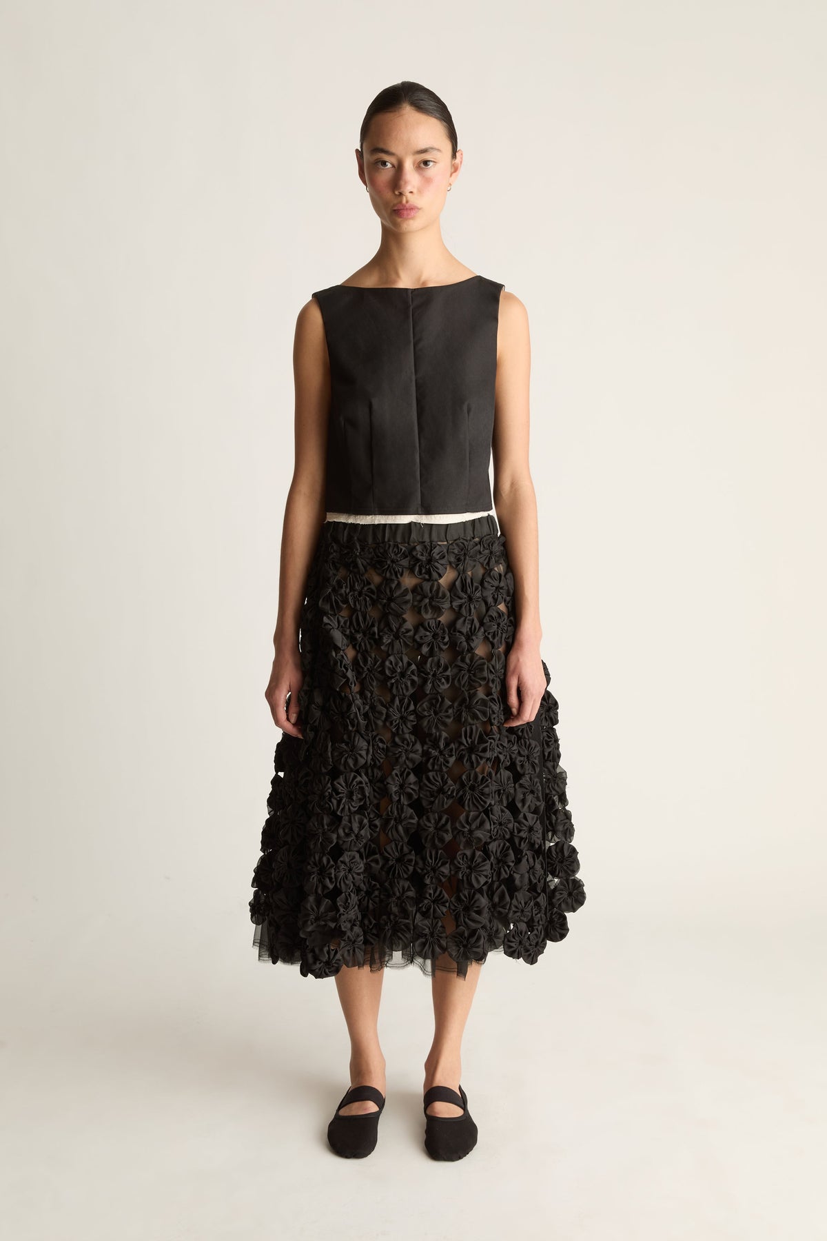 Front view of a woman wearing a black sleeveless top and black rosette midi skirt with tulle hem against a white studio background