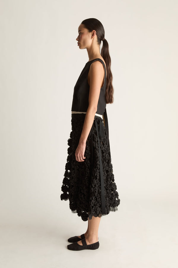 Side view of a woman with a ponytail in a black sleeveless top with cream waist trim and a black rosette midi skirt
